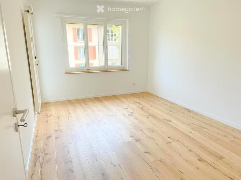 Empty room with light wooden flooring, white walls, and a large window with a view of a neighboring building.