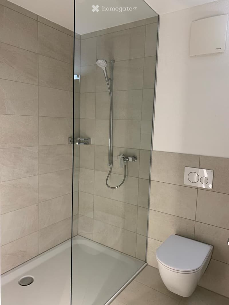 Modern bathroom with a glass-enclosed shower and a toilet. The shower features a handheld showerhead and a built-in seat. The walls and floor are tiled in a neutral beige color.