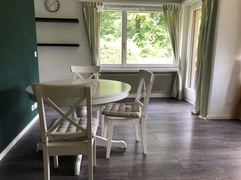 A dining room with a round white table, four white chairs, green walls, and a window overlooking the trees.