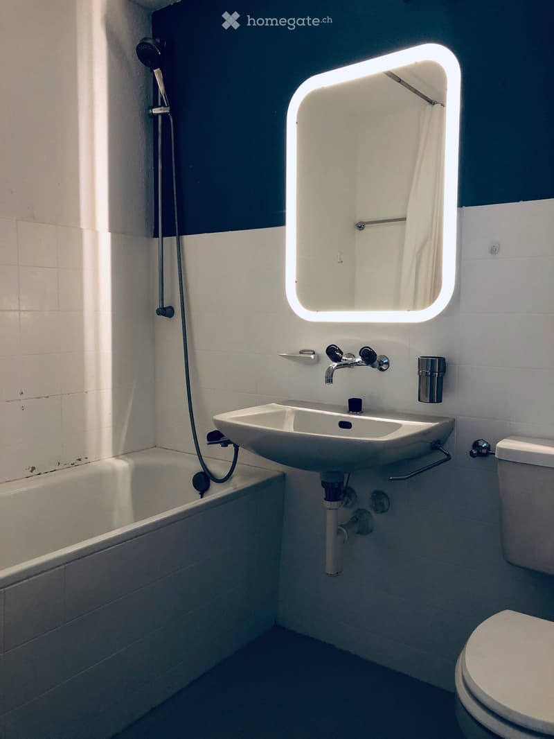 white tiled bathroom with bathtub, a white wall-mounted sink, a lit mirror above the sink, and a toilet with closed lid.