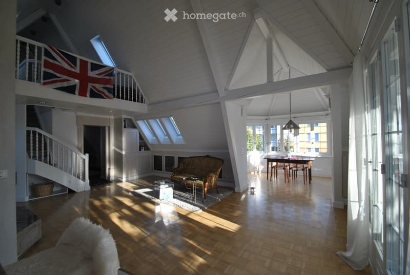 Bright, open-plan living and dining area with wooden floors, high ceilings, large windows, and skylights. Features a patterned sofa, coffee table, dining table with chairs, and a fluffy white rug.