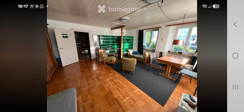 A cozy living room with wooden flooring, a grey sofa, a wooden dining table with chairs, and a small kitchen area with green tiles and cabinets.
