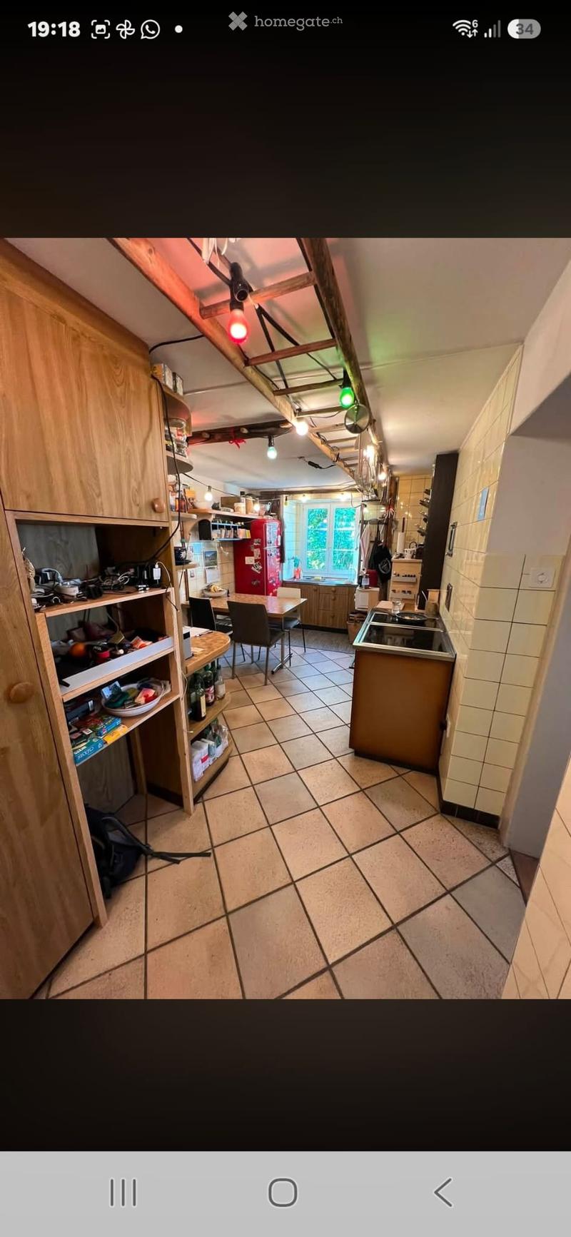 The kitchen features a tiled floor, wooden cabinets, a refrigerator, a sink, a stove, a dining table with chairs, and shelves with various items.