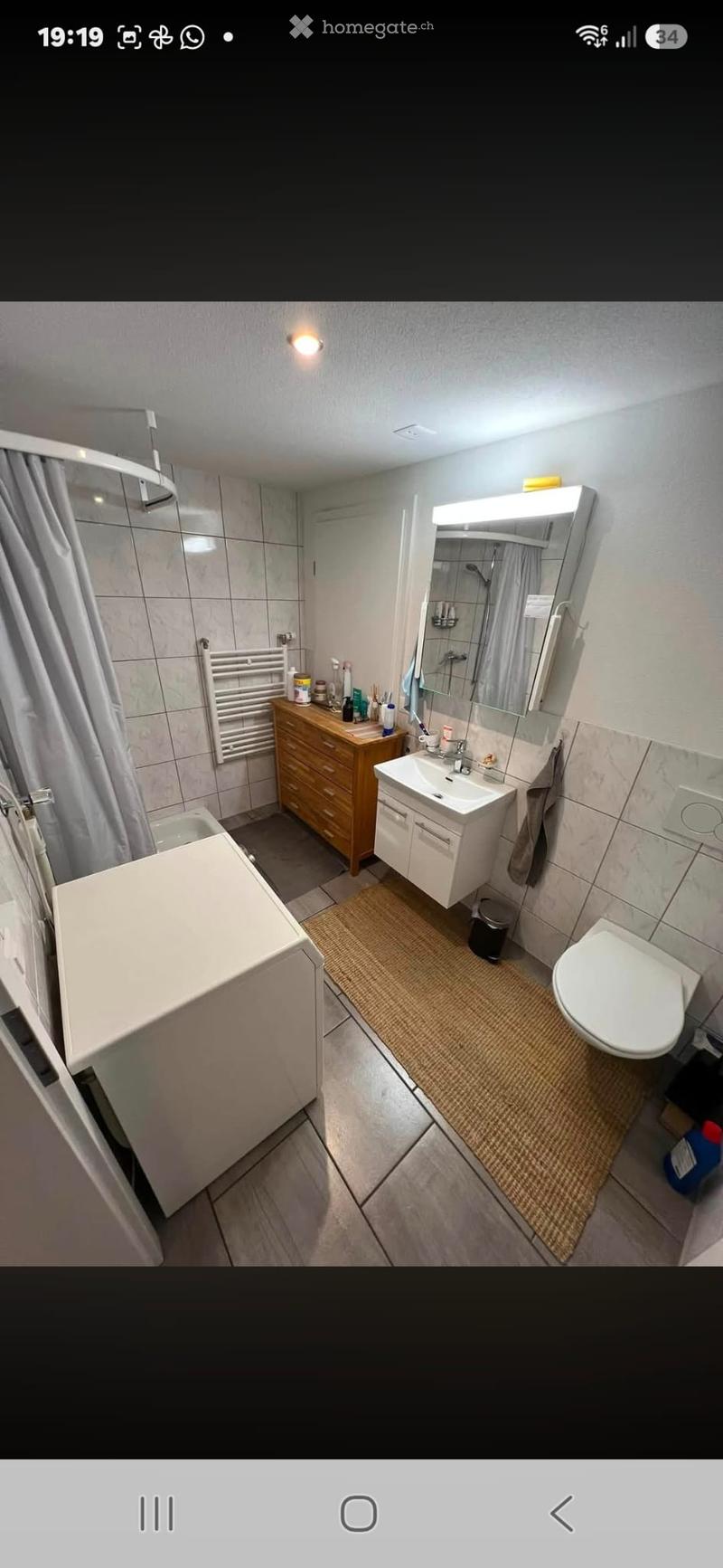 Modern bathroom with a white sink, toilet, mirror, wooden cabinet, and a shower area with a curtain. Includes a towel rack and a small rug on the floor.
