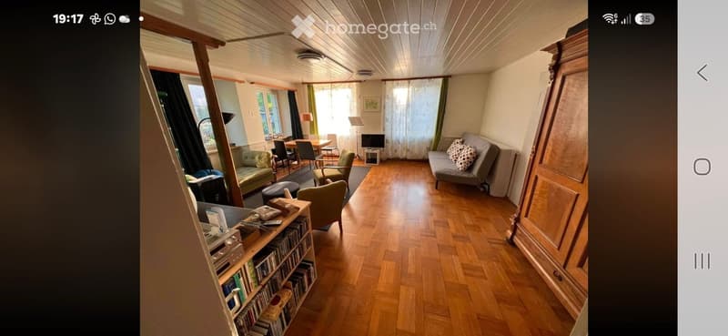 A cozy living room with wooden flooring, a grey sofa, a dining table with chairs, a bookshelf, and a television. The room is well-lit with natural light from large windows and has a warm, inviting atmosphere.