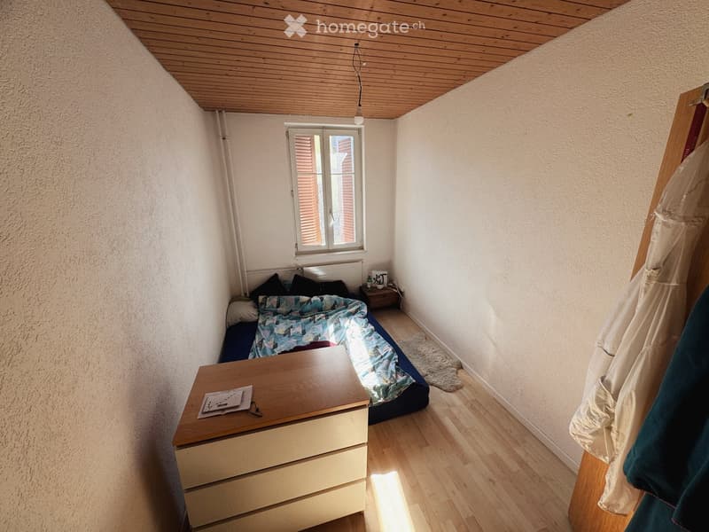 Cozy bedroom with wooden floor, slanted wooden ceiling, bed with colorful quilt, nightstand, small table, and radiator under window.