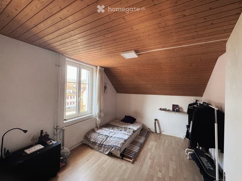 Cozy attic bedroom with wooden slanted ceiling, bed, nightstand, lamp, window with curtain, radiator, shelf, and clothing rack.