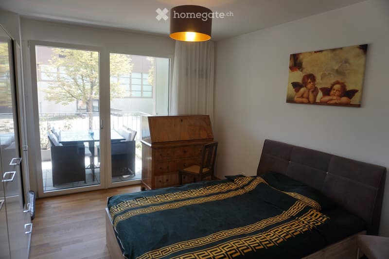 Modern bedroom with a large bed, dark green duvet, wooden floor, large window with balcony access, and a painting on the wall.
