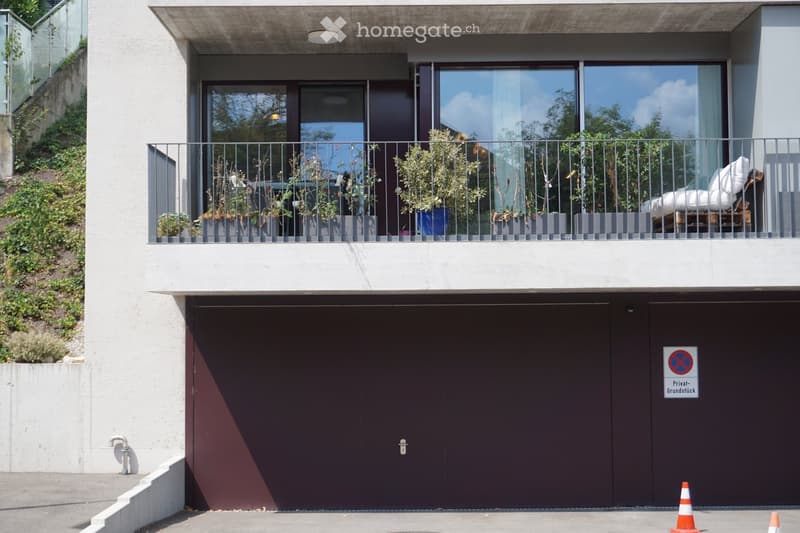 Modern two-story building with a large garage entrance, a balcony with potted plants and outdoor seating, and a raised ground floor.