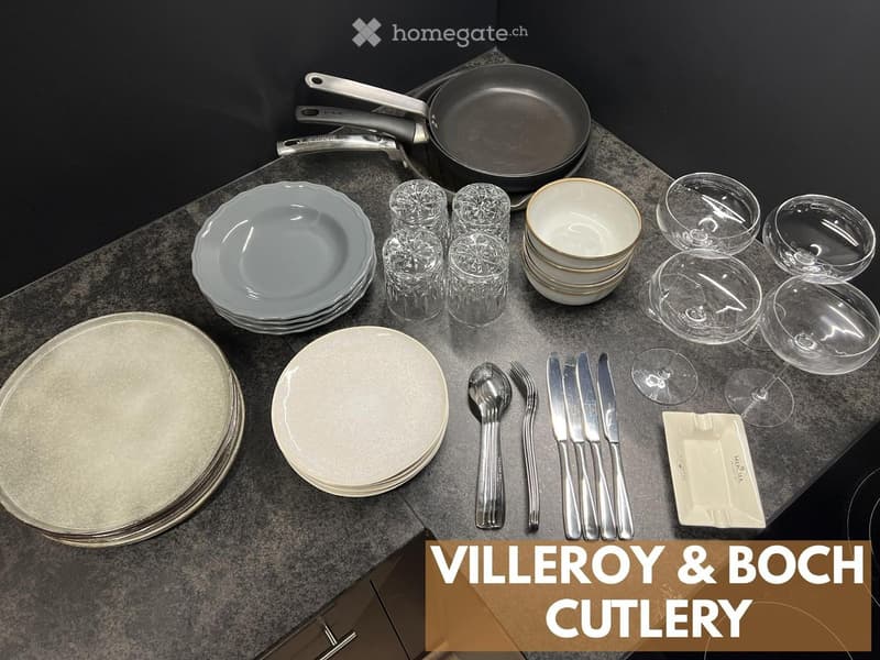 A kitchen countertop displaying a set of fine dining dishes and cutlery, including plates, bowls, wine glasses, and a frying pan.
