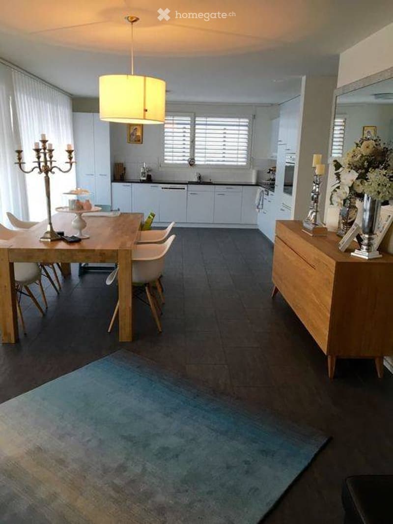 Bright, open-plan dining and kitchen area with modern white cabinets, a wooden dining table surrounded by white chairs, a candelabra, a large mirror, and a vase with flowers on a sideboard. The space features large windows with blinds and a hanging light