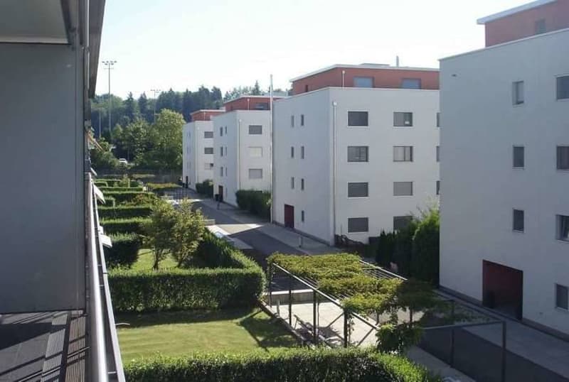 View from a balcony overlooking a landscaped garden area with trimmed hedges, small trees, and a pathway leading to a modern residential building complex.