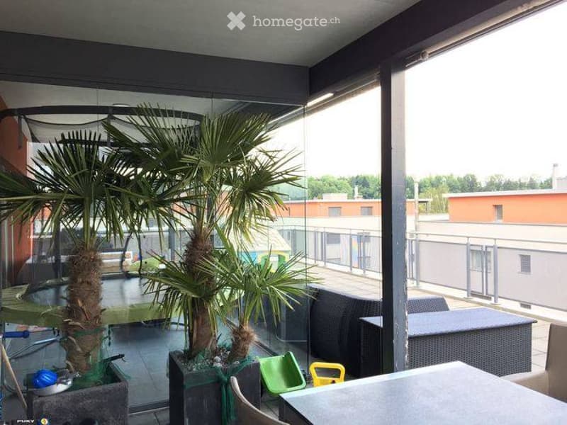 Modern balcony with large glass walls, two potted palm trees, outdoor furniture including black wicker chairs and tables, and a small green and yellow plastic chair.
