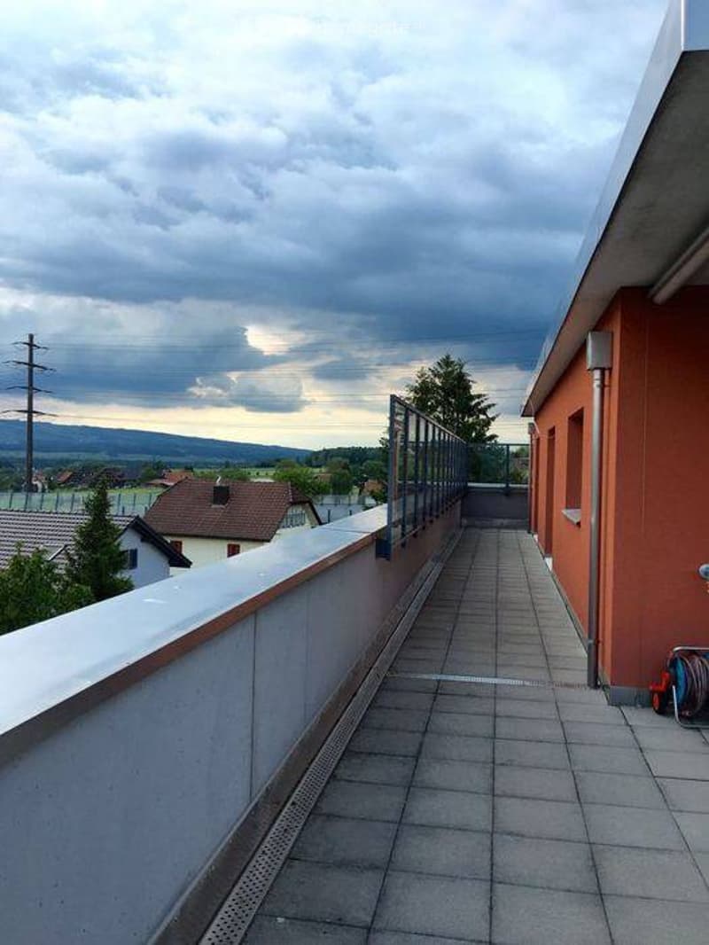 Balcony with tiled floor, white railing, and view of surrounding area and distant hills.