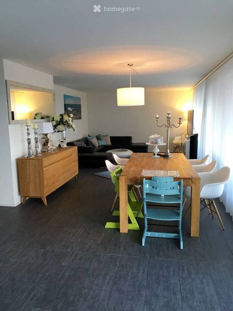 Modern living and dining area with a dark wood floor, a black sofa, a wooden dining table with chairs, a wooden cabinet, and decorative items including candles and a vase.