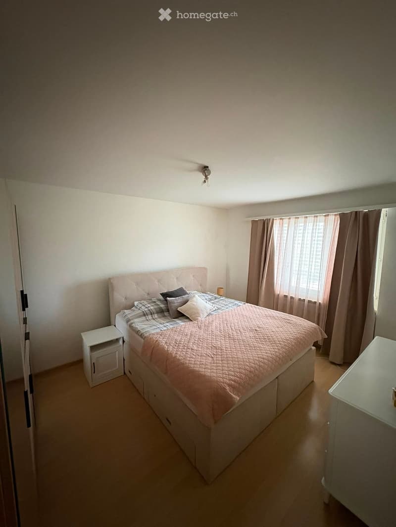 A well-lit bedroom with a large bed, nightstand, and a small table near the window with curtains.