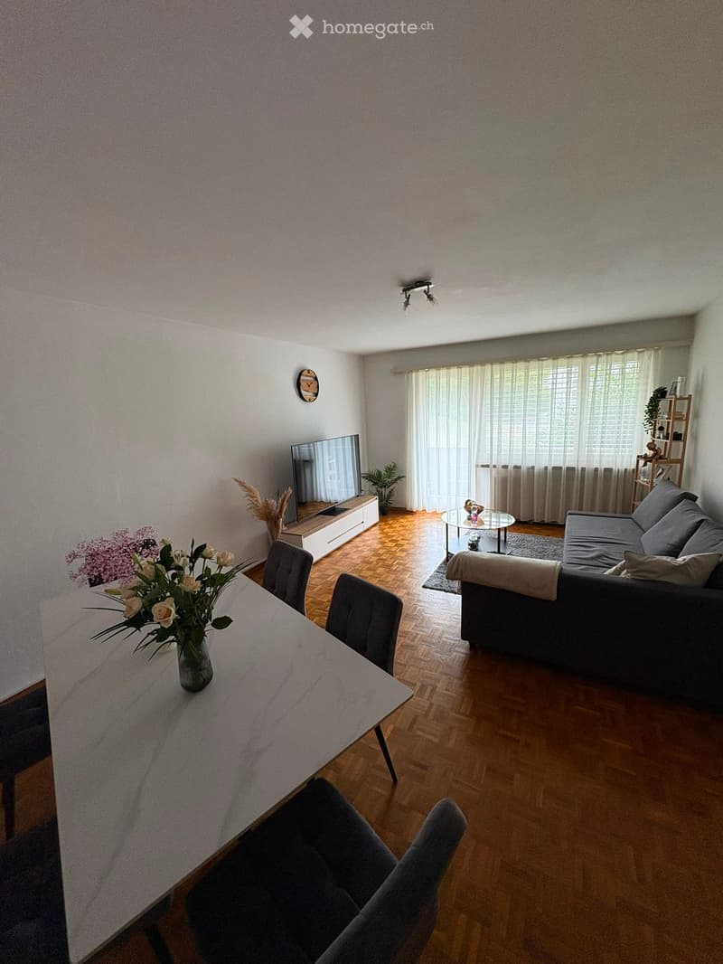 Modern living room with wooden flooring, large window with curtains, grey sofa, coffee table, dining table with chairs, TV, and decorative plants.