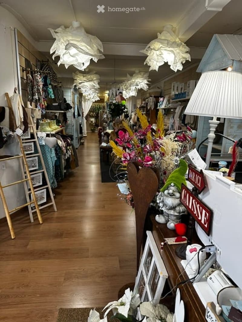 A well-lit, cozy retail or boutique space with wooden flooring, hanging floral lamps, shelves filled with clothing and accessories, and various decorative items including flower arrangements and picture frames.