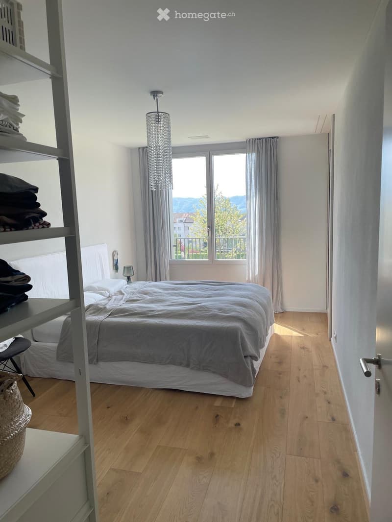 A modern bedroom with a wooden floor, a large bed with grey bedding, a chandelier, a shelf with folded clothes, and a window with curtains offering a scenic view.