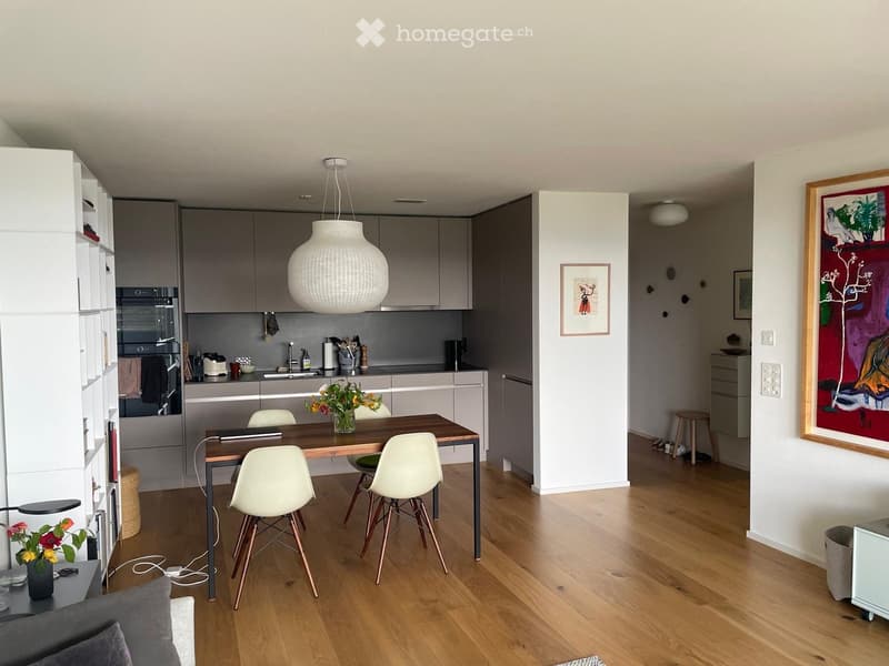 Modern kitchen and dining area with wooden flooring, white cabinets, stainless steel appliances, a round pendant light, and a dining table with chairs.