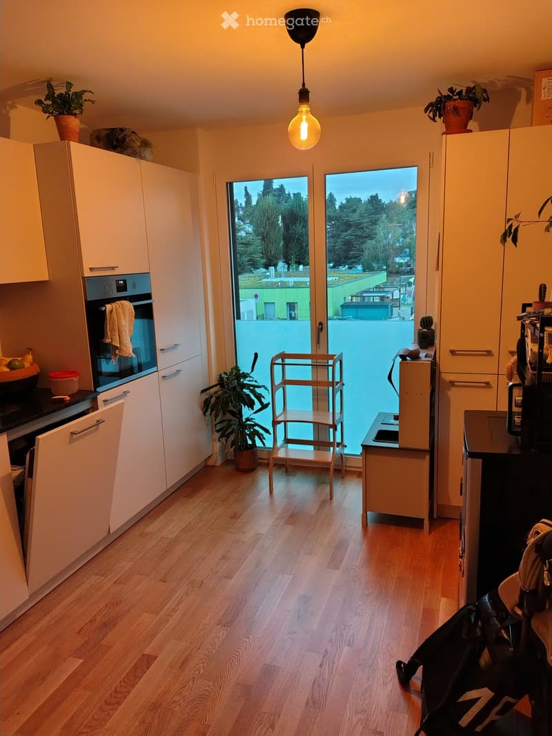 Modern kitchen with light wooden flooring, white cabinets, an oven, a refrigerator, a small table with a chair, and potted plants. Large windows offer a view of the outdoor area.