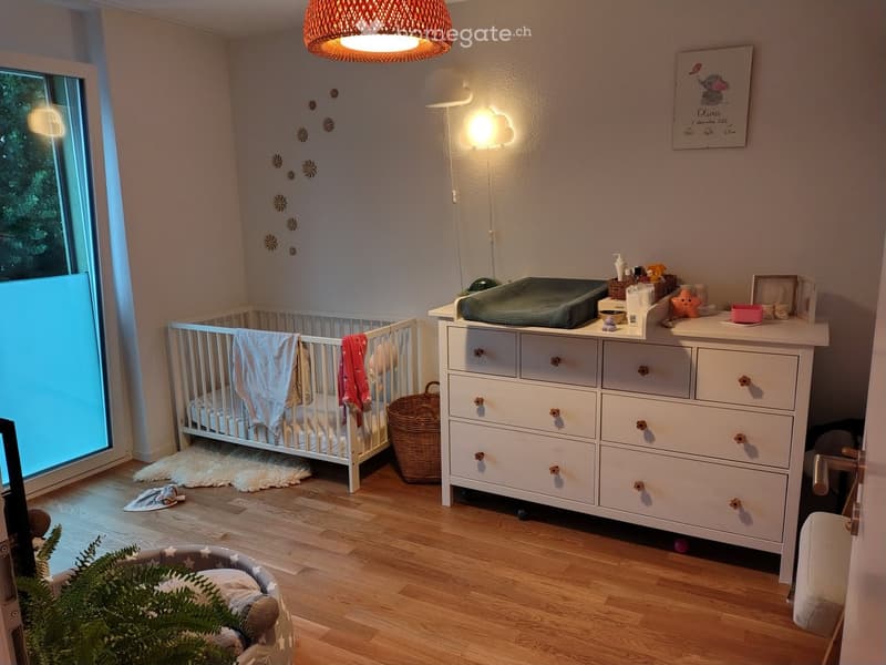 A well-lit nursery with a crib, dresser, and various baby items. The room features wooden flooring, a large window, and decorative wall stickers.