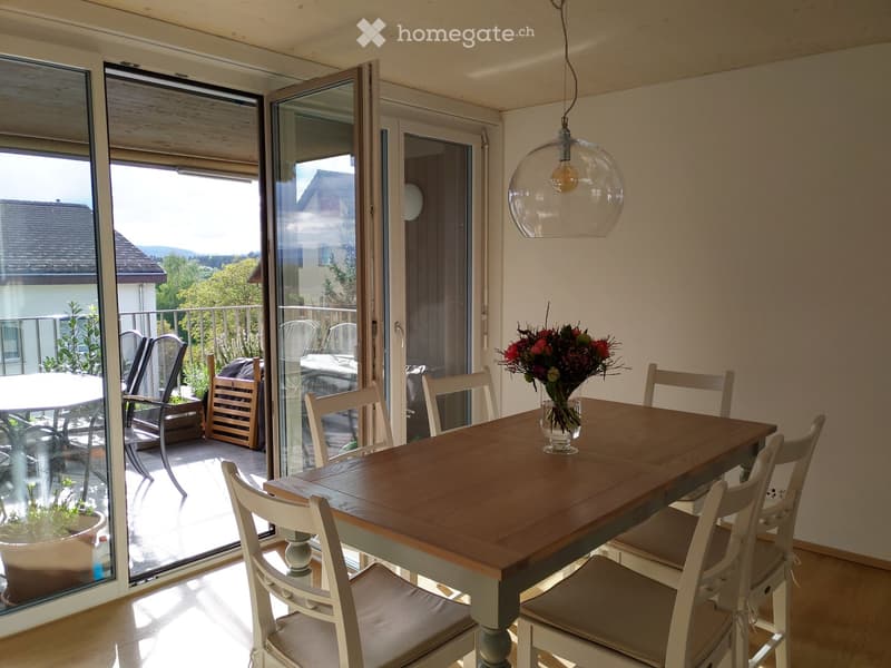 Bright dining room with a wooden table, white chairs, a flower vase, and a pendant light. Large sliding glass doors open to a balcony with outdoor furniture and a scenic view.