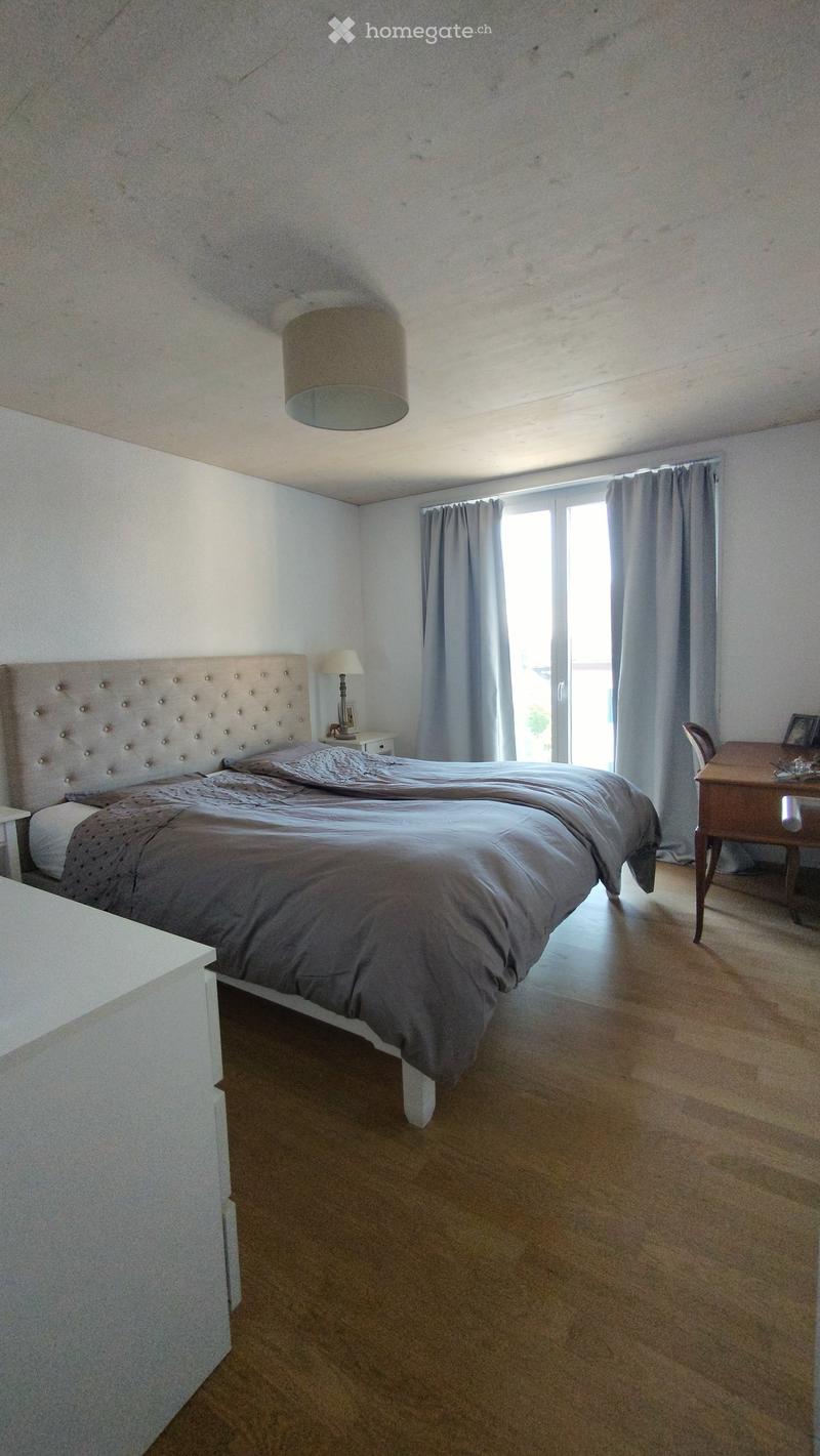 Bedroom with a large bed, nightstand, lamp, chair, and table near a large window with curtains.