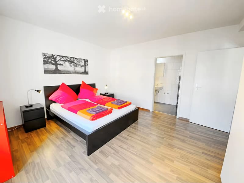 Bright bedroom with a double bed, colorful bedding, nightstands, and a lamp. A picture frame hangs on the wall, and a bathroom is visible through an open door.