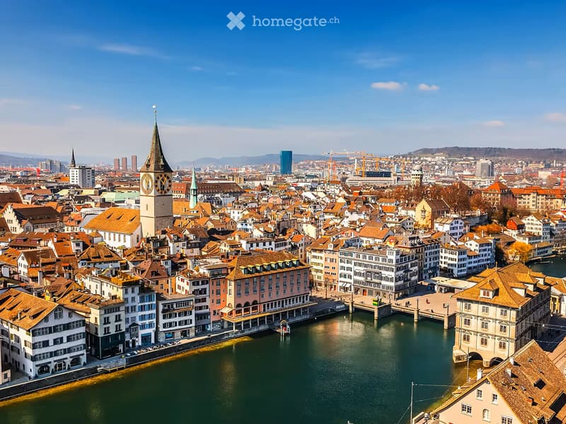 A panoramic view of a cityscape with a prominent clock tower, numerous buildings with red-tiled roofs, and a river running through the center.