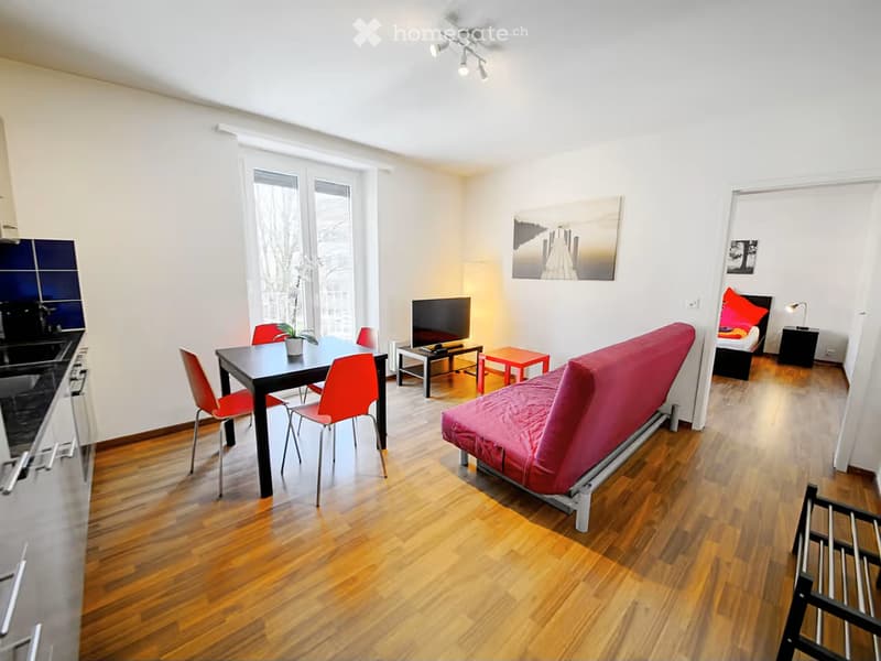 A modern, open-concept living and dining area with a red sofa, black dining table with red chairs, a television, and a small bedroom area with a bed and nightstand.