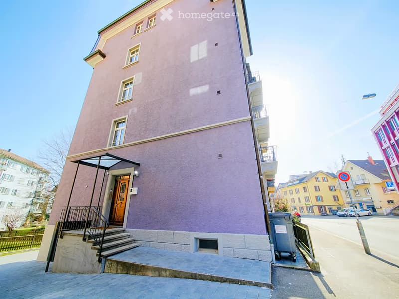 A multi-story residential building with a pink facade, white trim, and multiple windows. The entrance features a staircase leading to a wooden door, and there is a small balcony on the upper floor.