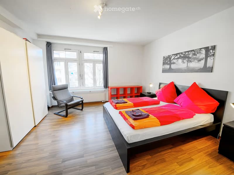 Bright bedroom with wooden flooring, large window, double bed with colorful bedding, red shelving unit, and a modern chair.