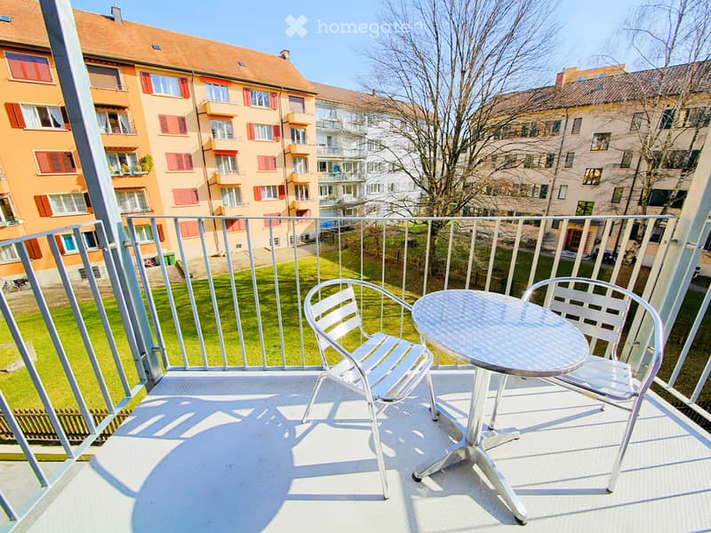 Balcony with a round metal table and two matching chairs, overlooking a green lawn and residential buildings.