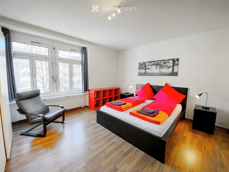 Modern bedroom with a large bed, colorful bedding, a red shelf, a grey chair, and a nightstand with a lamp.