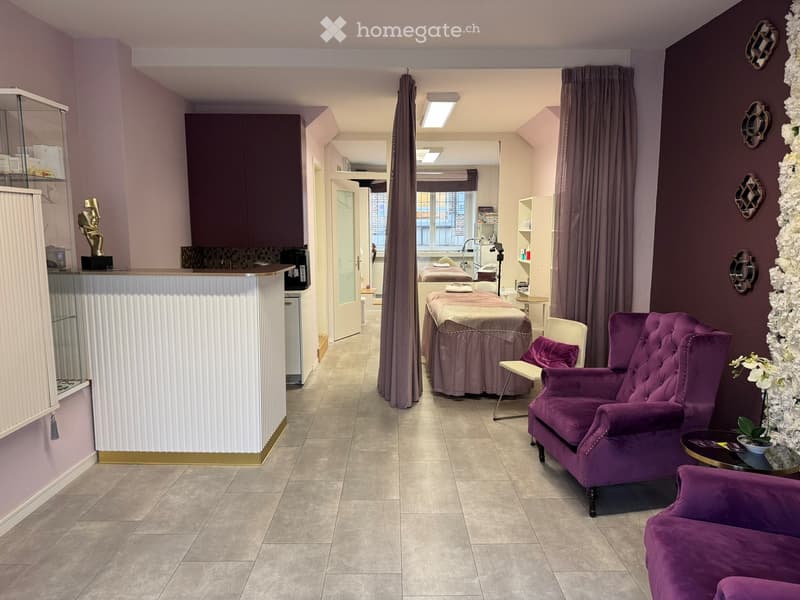 Well-lit spa or relaxation room with purple upholstered furniture, a massage table, decorative mirrors, and a reception area with a cabinet and statue.