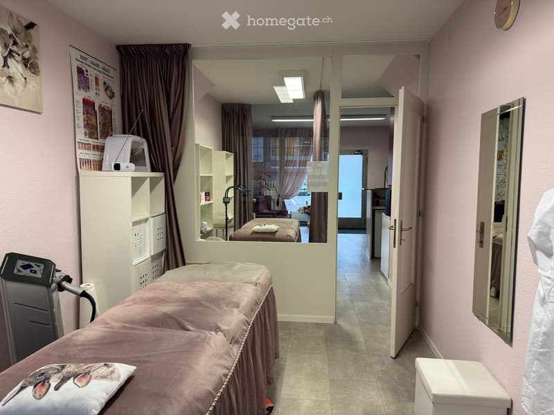 A well-lit, clean treatment room with a pink treatment table, a large mirror, a cabinet, shelves, and a lamp. The room features light pink walls and a tiled floor.