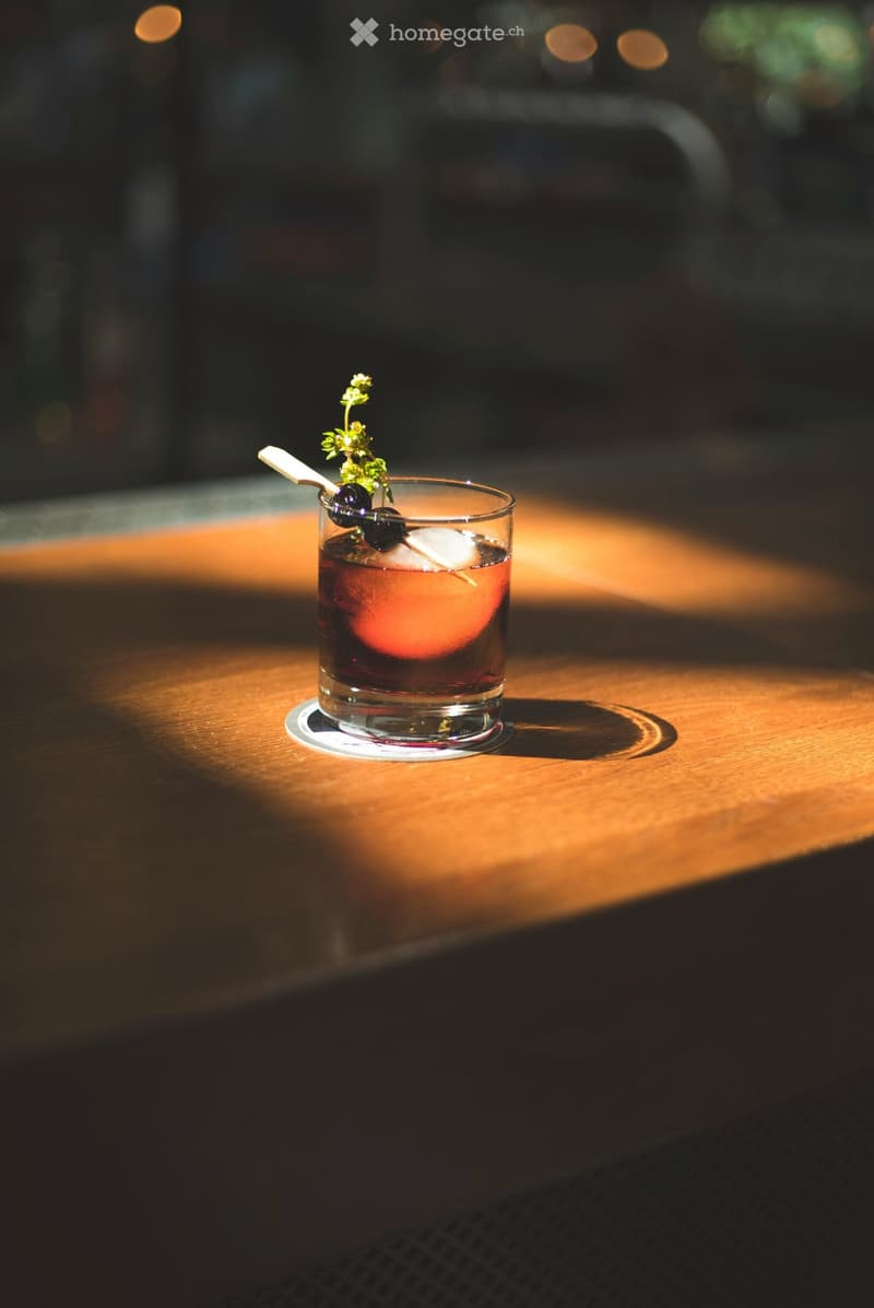 A cocktail in a short glass with ice, olives, and a herb garnish on a wooden surface.