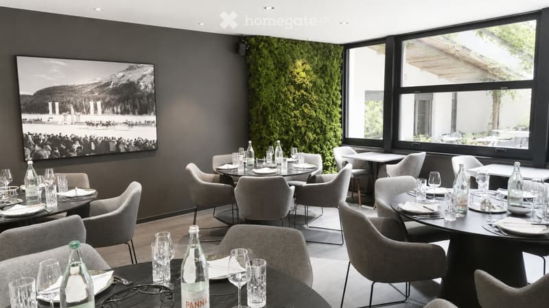 Modern restaurant interior with round tables, grey chairs, and large windows overlooking an outdoor area. Tables are set with plates, glasses, and bottles.