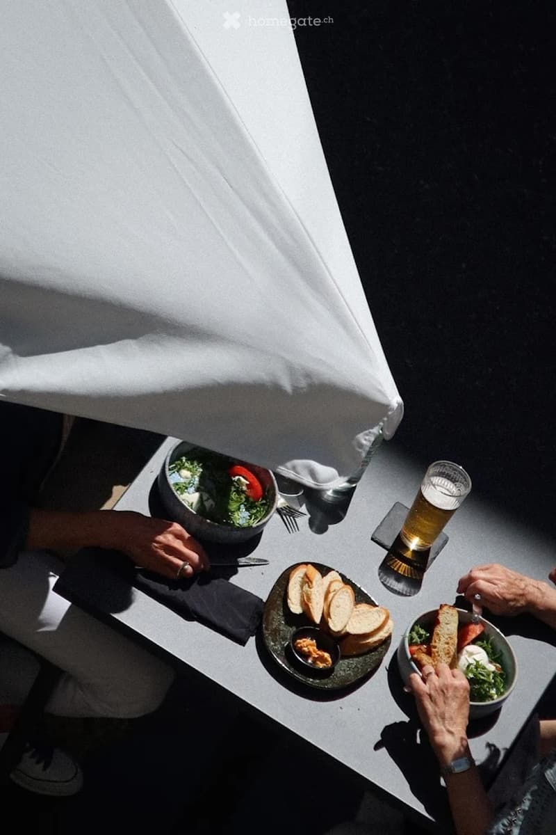 An outdoor dining setup with a table covered by a white umbrella, featuring plates of food, a bowl of salad, and a glass of beer.