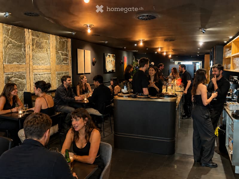 A modern restaurant interior with patrons seated at tables and standing near the bar, featuring dark wooden tables, chairs, and a central bar area with stools. The decor includes stone and wood paneling, modern lighting, and artwork on the walls.