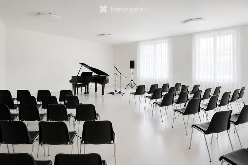 A spacious, well-lit room with white walls and a polished floor, featuring rows of black chairs arranged facing a grand piano, a microphone stand, and a speaker.