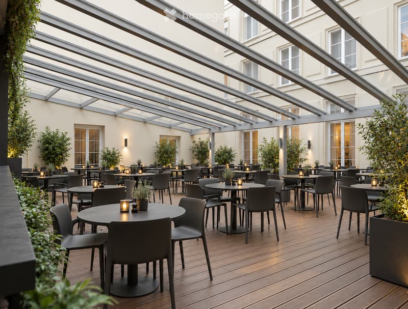 An outdoor dining area with wooden flooring, round tables, and chairs arranged under a modern metal pergola. Potted plants and small candle holders are placed on the tables.