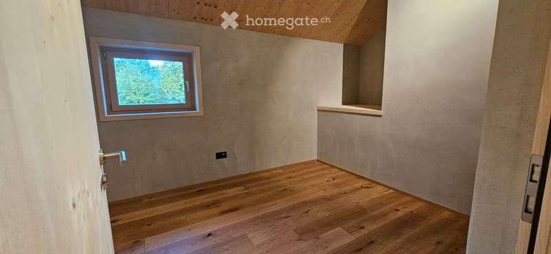 Empty room with wooden flooring, white walls, and a single window with a view of greenery. The room features a small built-in shelf and a door with a silver handle.