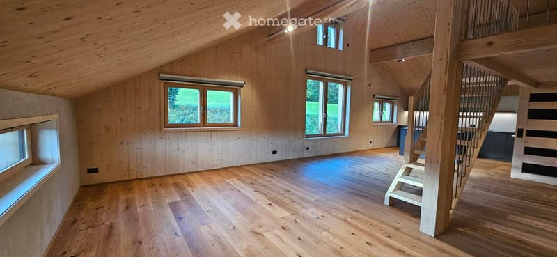 Bright, open loft space with wooden walls and floors, large windows, and a staircase leading to an upper level.