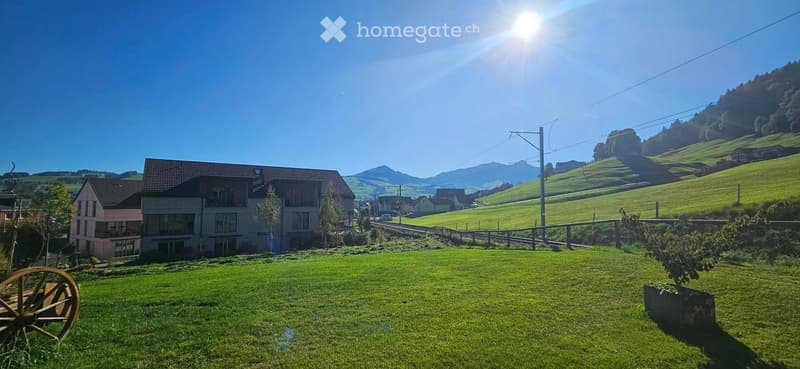 A large, well-maintained lawn in front of a two-story house with a red-tiled roof, surrounded by rolling green hills and distant mountains under a clear blue sky.