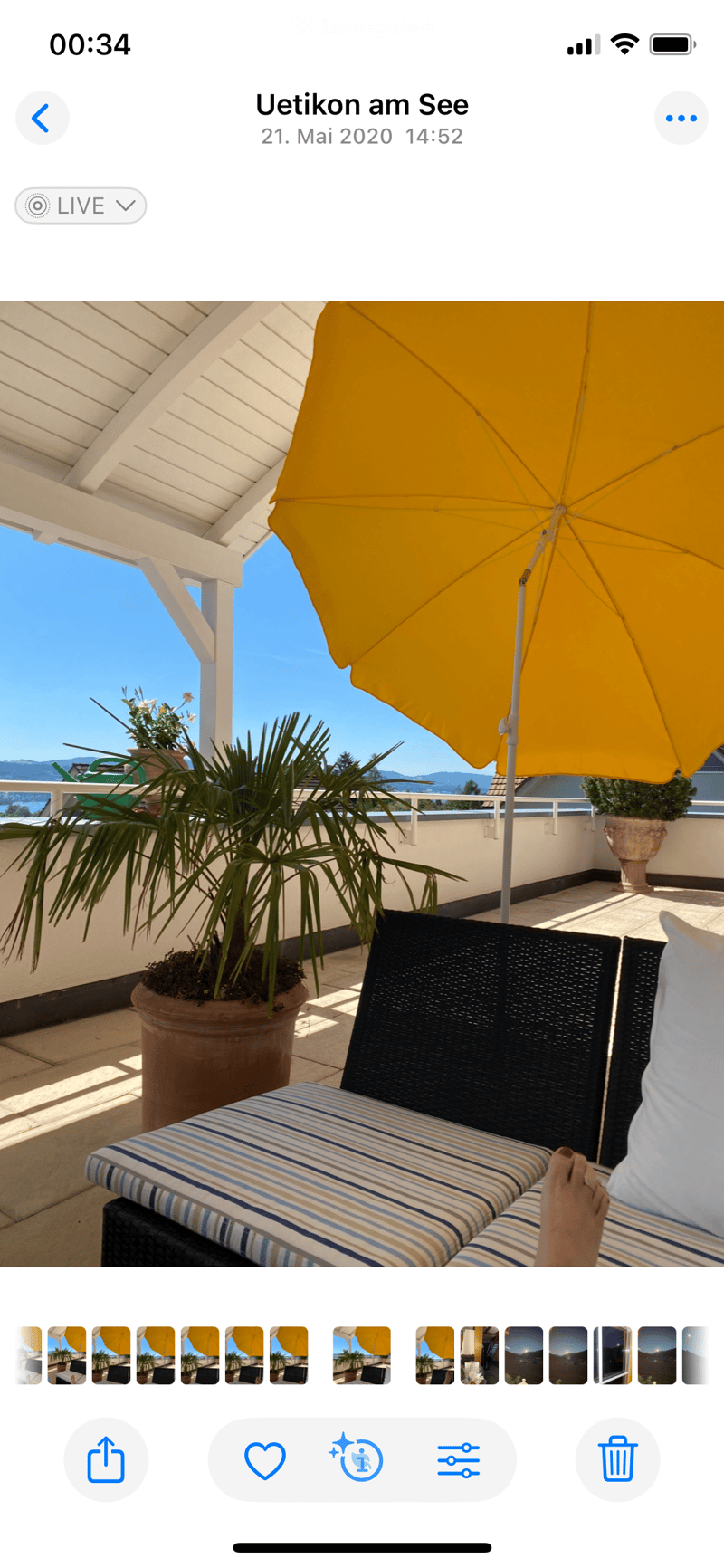 A sunny balcony with a large yellow umbrella, potted plants, and striped lounge chairs, overlooking a scenic view.