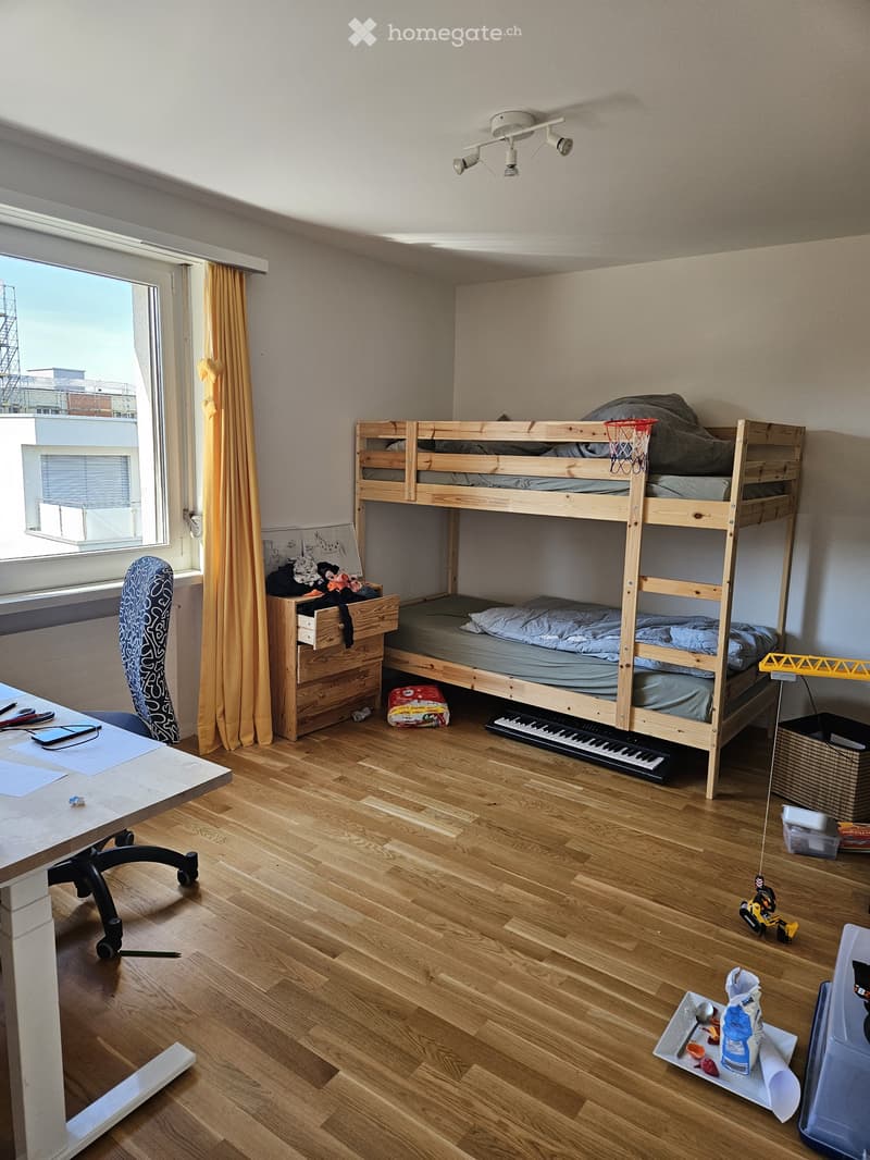 A small, multi-functional room with a lofted bed, desk, and various items including a keyboard, toy car, and a mirror. The room features wooden flooring and a window with a view of a construction site.