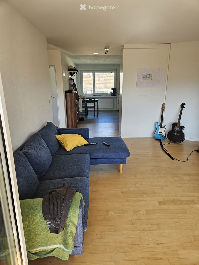 A compact, furnished room with a sofa, guitar on the wall, a small table, and a window providing natural light.