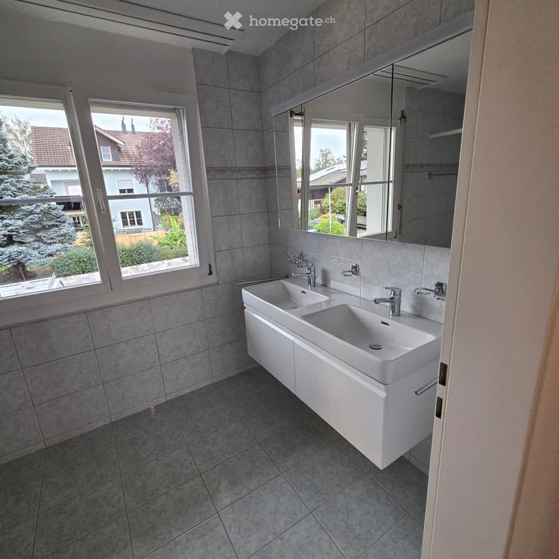 Modern bathroom with grey tiled walls and floor, double sink with modern faucets, large mirror, and large windows offering a view of the garden and neighboring house.